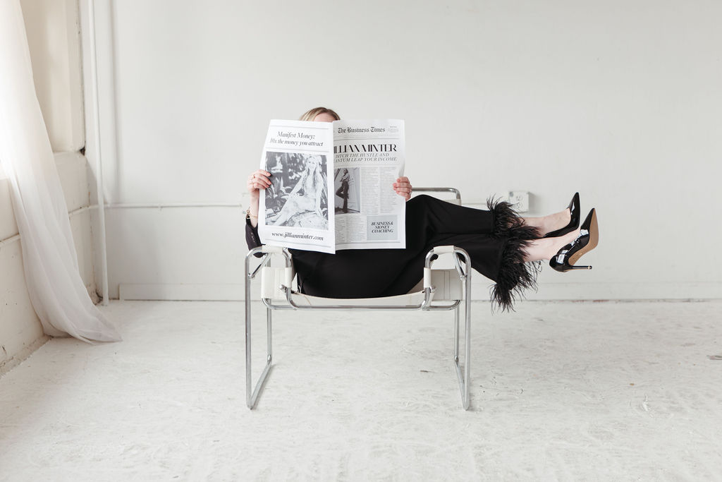 Here’s an alt text description for your image: Jillian, a business and money coach, lounges in a modern white and chrome chair with her legs crossed, wearing a black outfit with feathered cuffs and glossy black heels. She holds up a newspaper titled *The Business Times* with her name, "Jillian Minter," featured prominently, along with the phrase "Manifest Money: It’s the money you attract" and her website URL. The minimalist, airy setting and her poised posture convey confidence, financial success, and abundance.