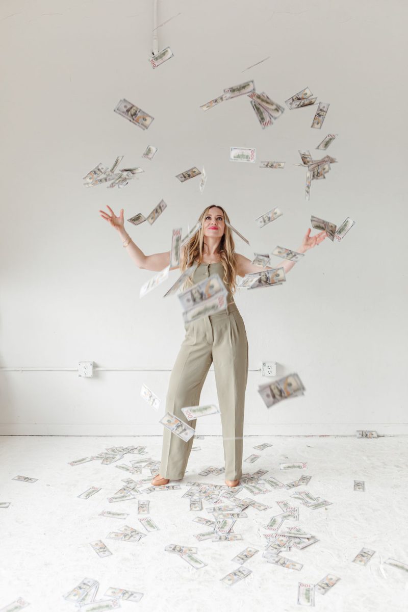 jillianminter-86 Here’s an alt text description for your image: Jillian, a business and money coach, stands in a minimalist white room, joyfully tossing stacks of cash into the air. She wears a stylish sage green jumpsuit, her arms open wide as the money rains down around her. The image exudes abundance, financial empowerment, and a mindset of wealth attraction.