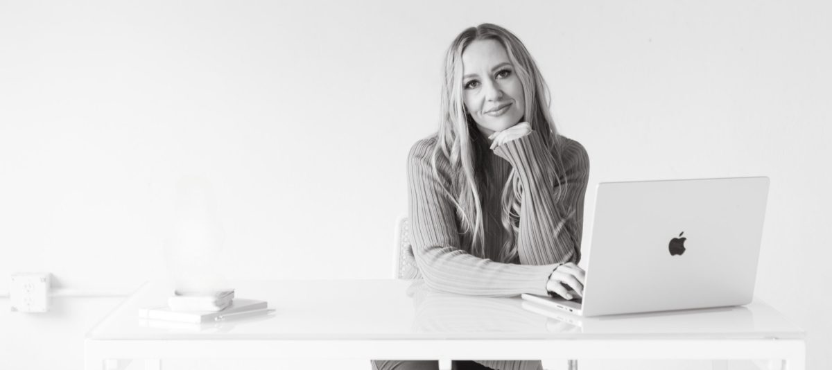 Black and white photo of Jillian, a money and business coach, sitting at a white desk with a MacBook in front of her. She’s resting her chin on her hand and smiling gently, wearing a ribbed sweater with long sleeves. The minimalist workspace includes a small stack of books and a pen on the desk.