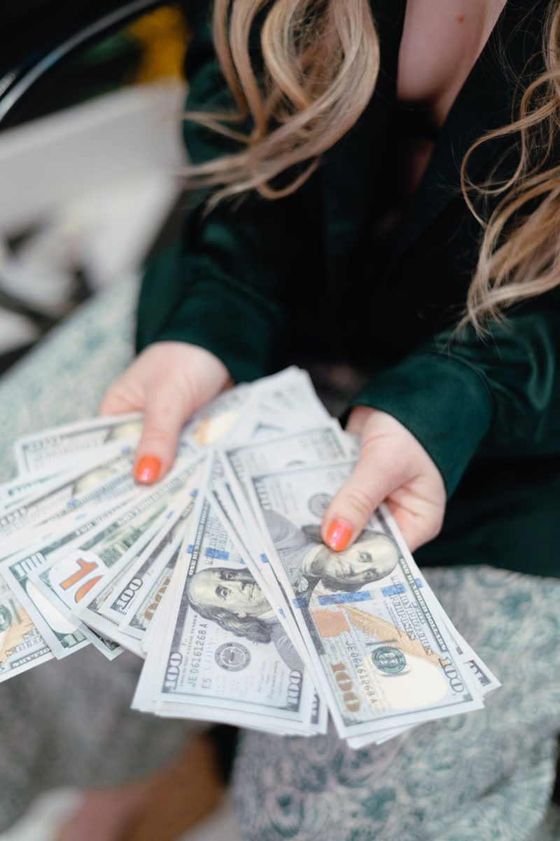 HVZL0938 Jillian, a money coach, holds a fan of $100 bills in both hands, her nails painted coral and her long blonde hair flowing over a dark green blouse. The image captures themes of wealth, empowerment, and financial confidence, aligning with her coaching message.