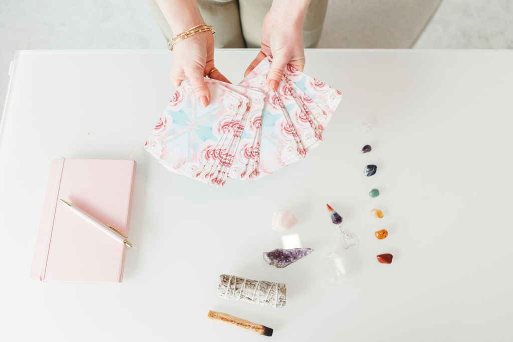 Jillian, a money coach expanding into spirituality, Reiki, and astrology, fans out a deck of pastel tarot or oracle cards over a white desk. The setup includes a pink journal with a pen, a row of colorful crystals, sage, and palo santo, highlighting a serene and intentional manifesting practice.
