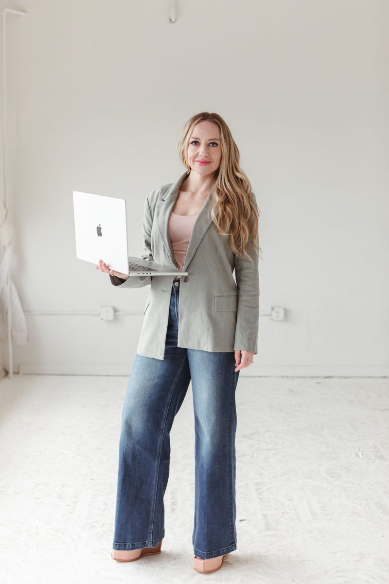 jillianminter-27 Jillian, a money coach, stands confidently in a bright, minimalist studio holding an open laptop. She’s wearing a sage green blazer, blush pink top, wide-leg jeans, and platform shoes, radiating approachability and professionalism.