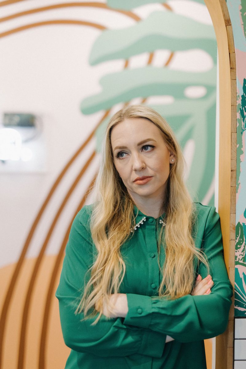 Jillian Minter, money mindset coach, stands with arms crossed and an exasperated expression, visibly frustrated by common money blocks, wearing a green blouse in front of a colorful abstract background.