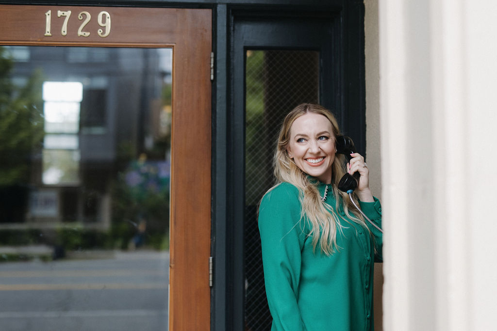 Jillian Minter, money mindset and manifestation coach, smiles brightly while holding a vintage-style black phone outside a building marked 1729, wearing a vibrant green blouse.