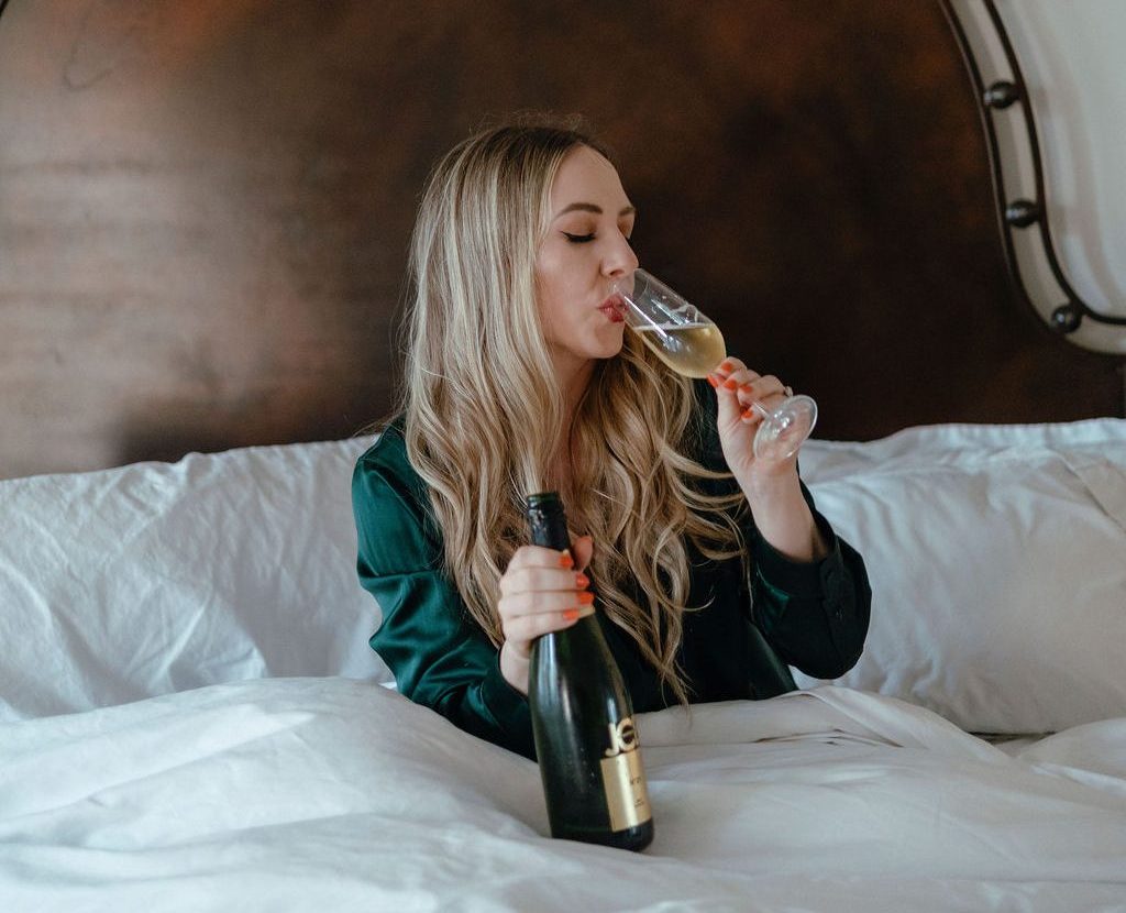 jillian-minter-money-coach-celebrating-success Jillian Minter enjoys a glass of champagne in bed, wearing dark green satin pajamas. With a champagne bottle in hand and a calm expression, the scene conveys a relaxed, luxurious moment of celebration.