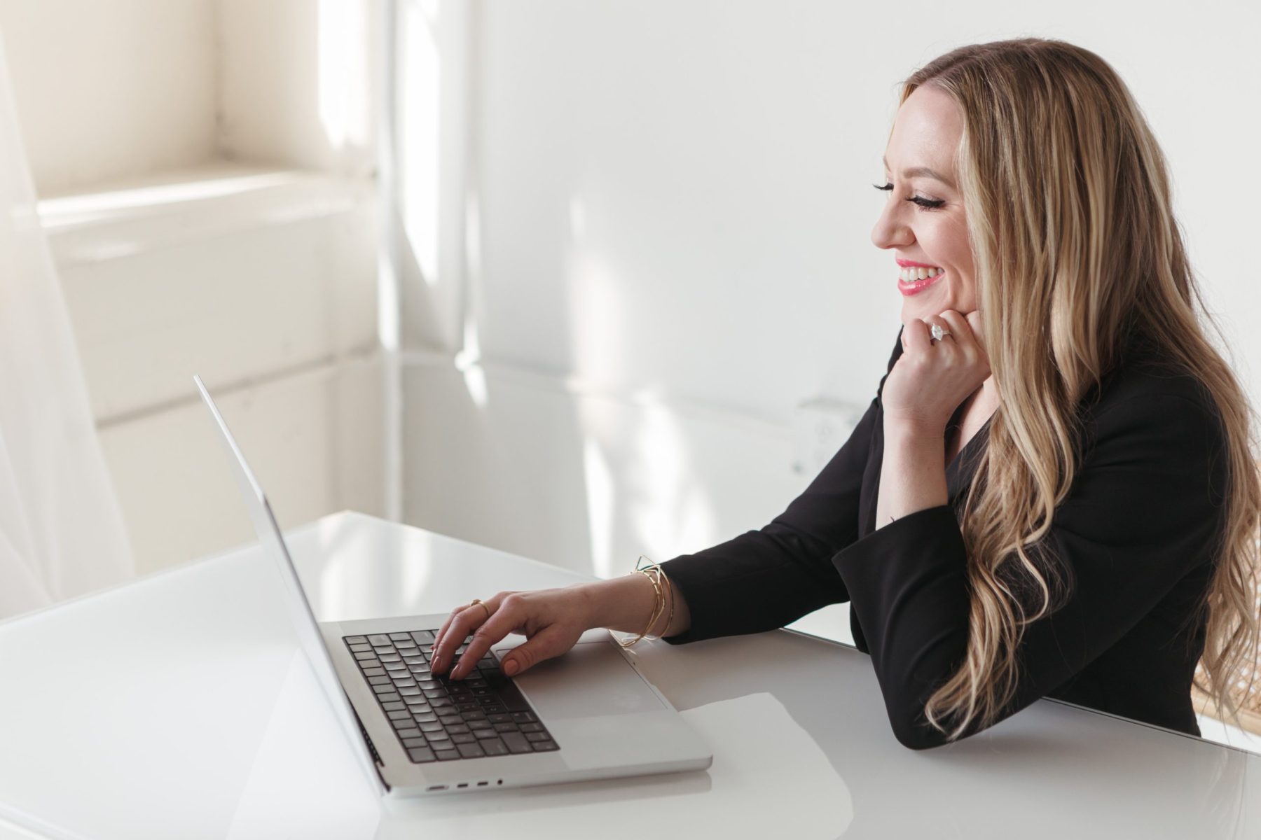 Jillian Minter, money mindset coach, smiles while working on her laptop at a sleek white desk, dressed in a black blazer in a sunlit office.