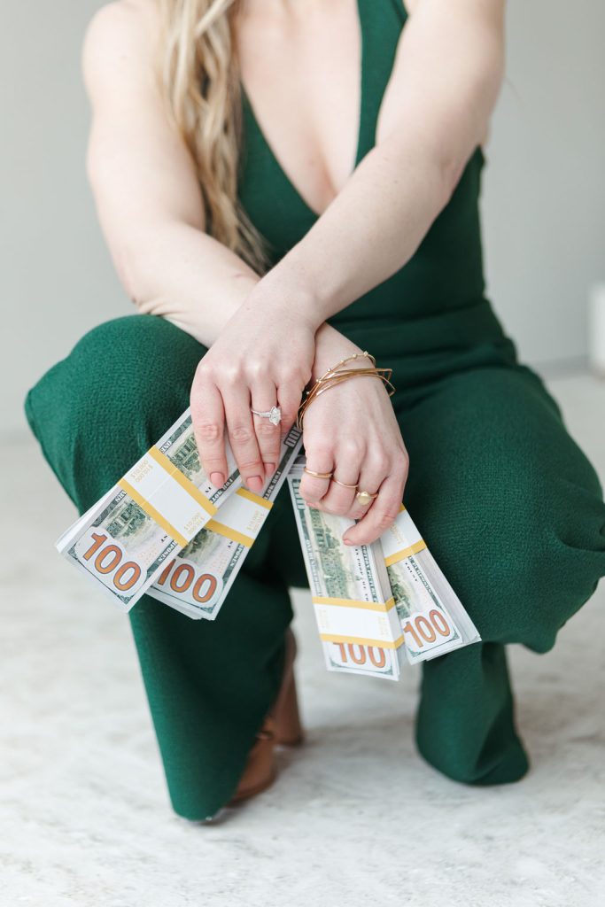 Close-up of Jillian Minter crouching in a green jumpsuit, holding large stacks of $100 bills in both hands, wearing gold and diamond jewelry.