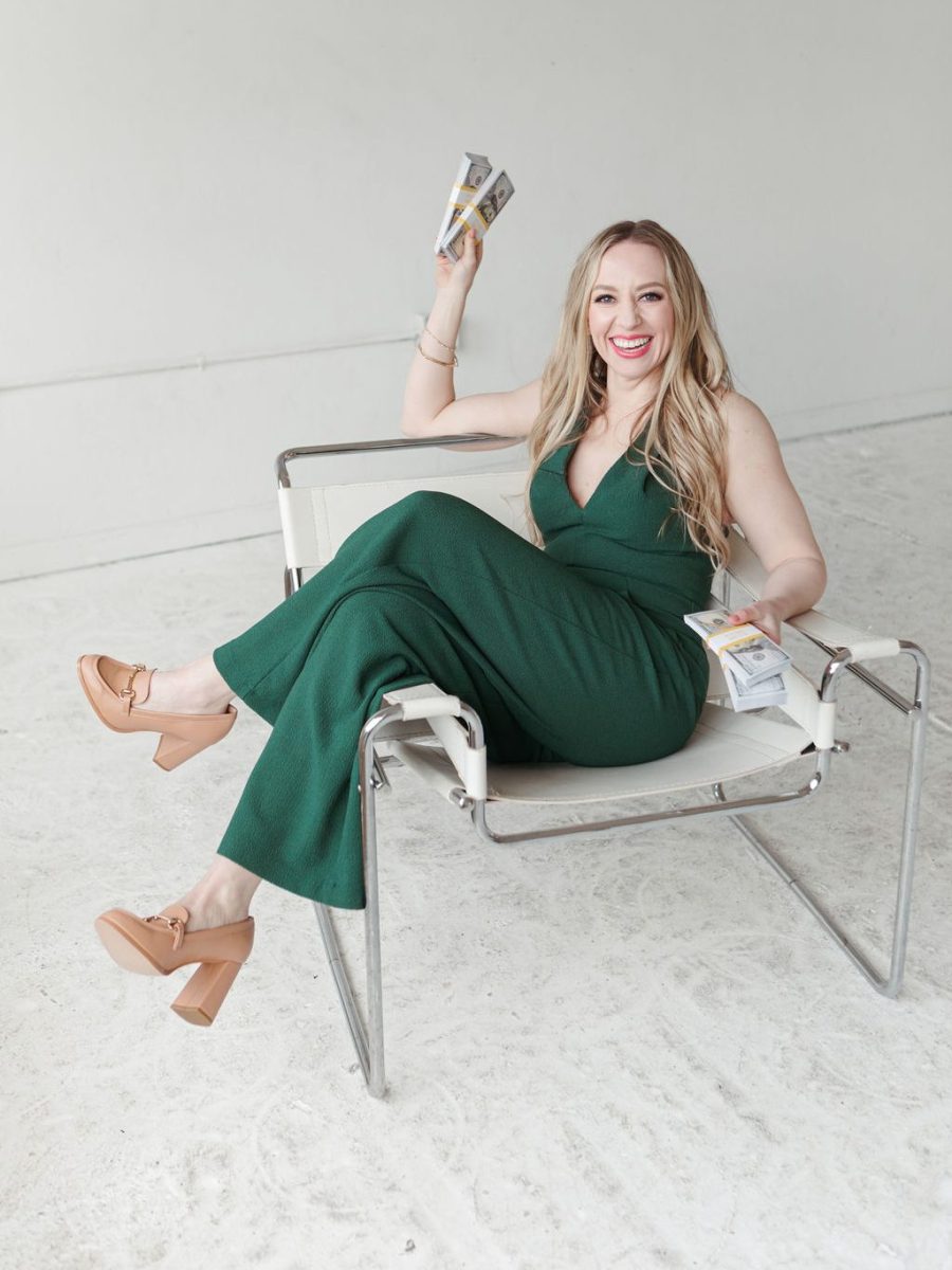 Jillian Minter sitting confidently in a modern white chair, wearing a green jumpsuit and tan heels, smiling brightly while holding thick stacks of $100 bills in both hands.