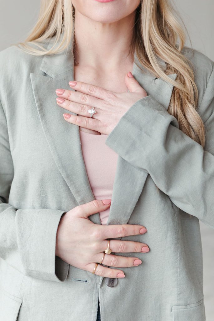A close-up of Jillian Minter placing one hand over her heart and one over her stomach, wearing a pale green blazer and soft pink top — a gesture of grounding, gratitude, and inner alignment.