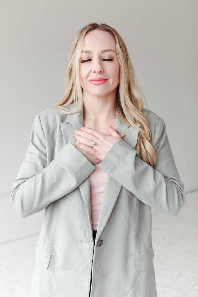 Jillian Minter, money mindset coach, stands with her eyes closed and hands over her heart, wearing a light sage blazer in a moment of calm alignment and gratitude.