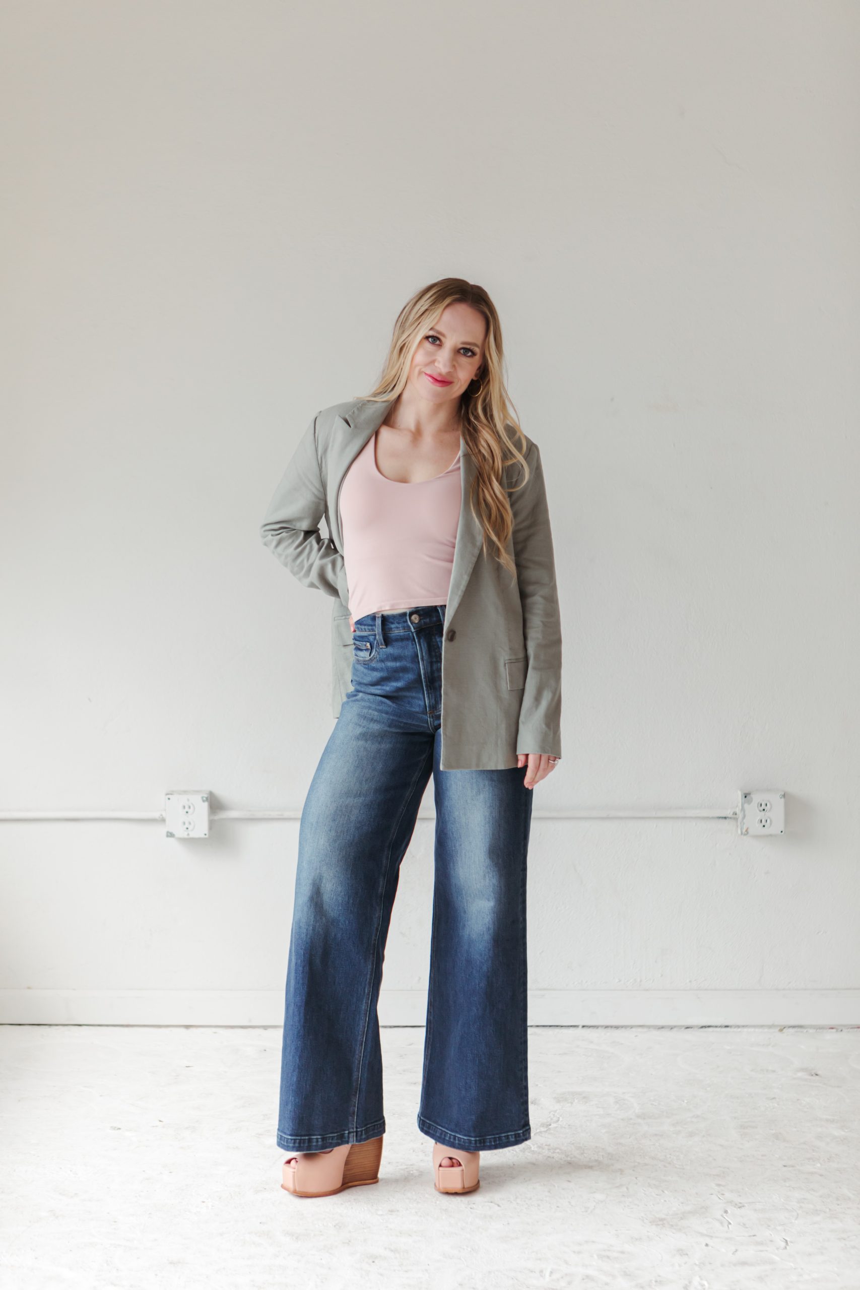 jillian-minter-money-mindset-coach-confident-personal-brand Jillian Minter stands confidently against a neutral wall wearing a light sage blazer, blush pink tank top, wide-leg jeans, and nude wedge heels, smiling with a relaxed and self-assured pose.