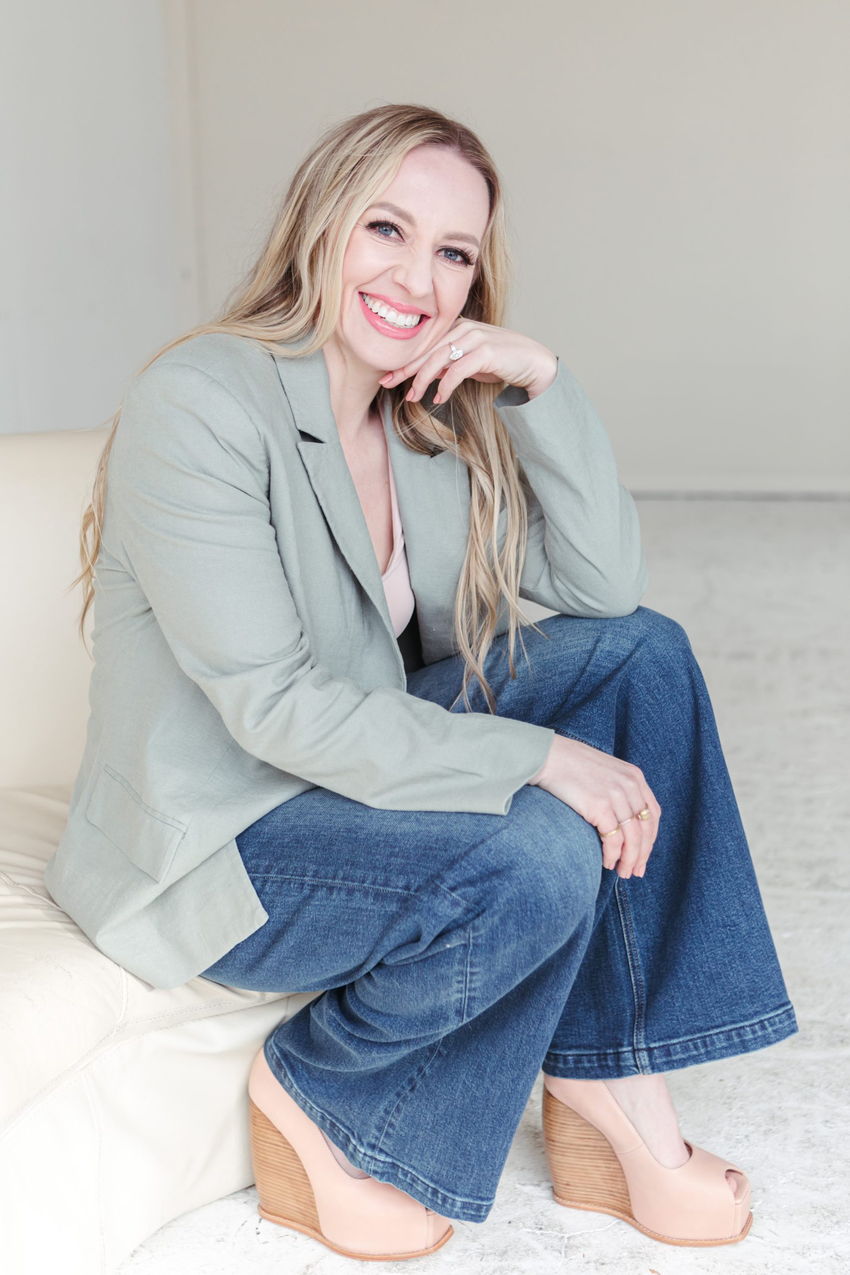 Jillian_Minter_seated_on_couch Jillian Minter, a business and money coach, sits sideways on a cream sofa, smiling brightly. She's wearing a sage green blazer, wide-leg jeans, and nude platform heels, radiating a confident and approachable vibe.