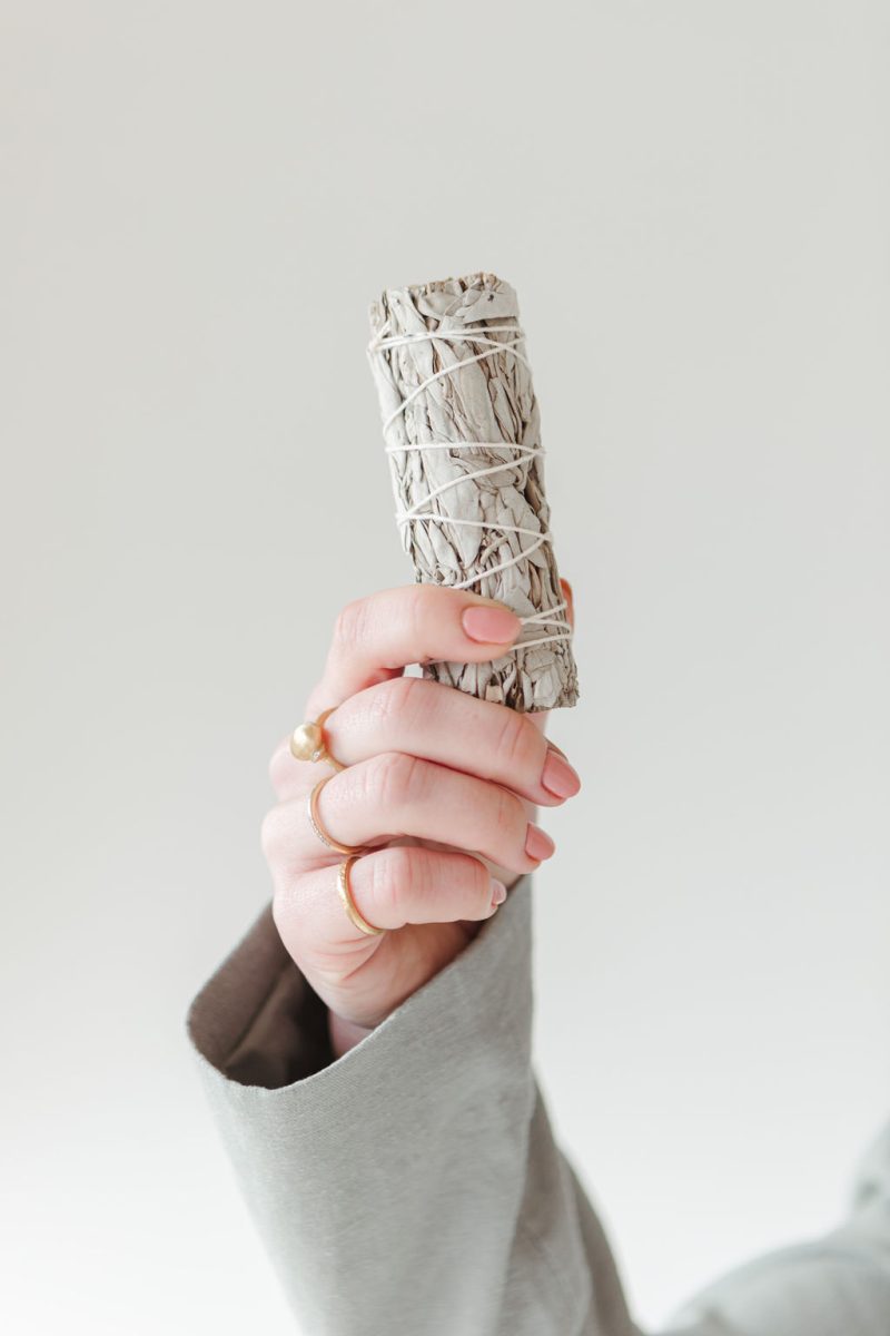 A hand wearing gold rings holds a wrapped sage bundle upright against a neutral background, symbolizing energetic clearing and intention-setting.