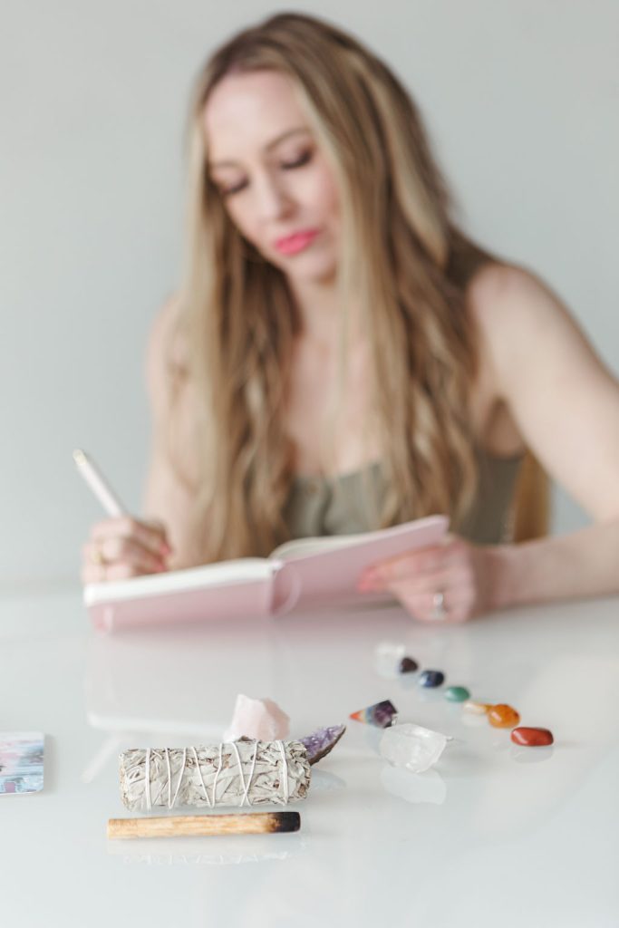 Jillian Minter, slightly out of focus in the background, journals in a pink notebook while in the foreground sits a sage bundle, palo santo, crystals, and chakra stones arranged on a white desk.