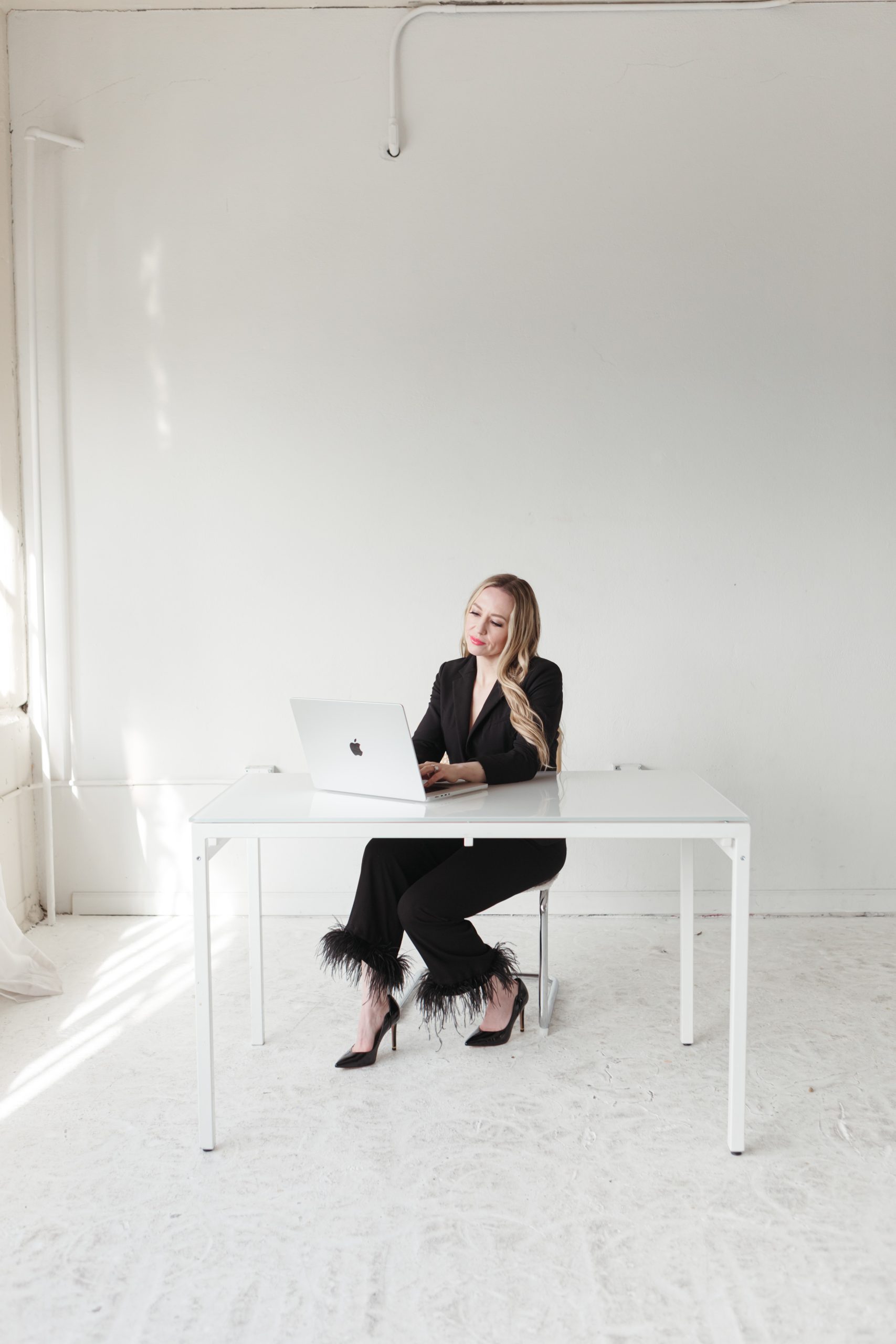 jillianminter-143 Jillian, a money coach, stands confidently in a bright, minimalist studio holding an open laptop. She’s wearing a sage green blazer, blush pink top, wide-leg jeans, and platform shoes, radiating approachability and professionalism.